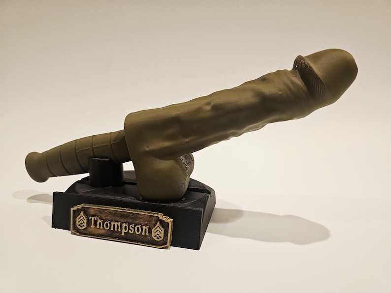 May include: A khaki-colored sculpture of a phallus mounted on a black base. The base features a gold-colored nameplate with the name "Thompson" and military insignia. The sculpture is detailed and realistic.