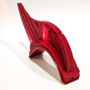 Op de afbeelding: Een rood kunststof onderdeel met een gebogen vorm en ingewikkelde details. Het onderdeel heeft een gladde, glanzende afwerking en lijkt een onderdeel te zijn van een groter apparaat of machine.