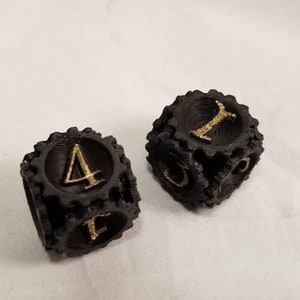 May include: Two black dice with gold numbers. The dice are shaped like gears and have a textured surface.