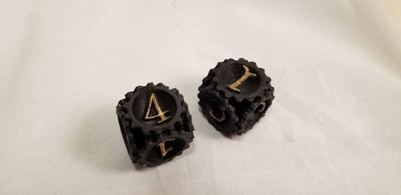 May include: Two black dice with gold numbers. The dice are shaped like gears and have a textured surface.
