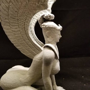 May include: A white, 3D printed sculpture of a winged woman with a crown, kneeling on all fours. The sculpture is detailed and has a textured surface.