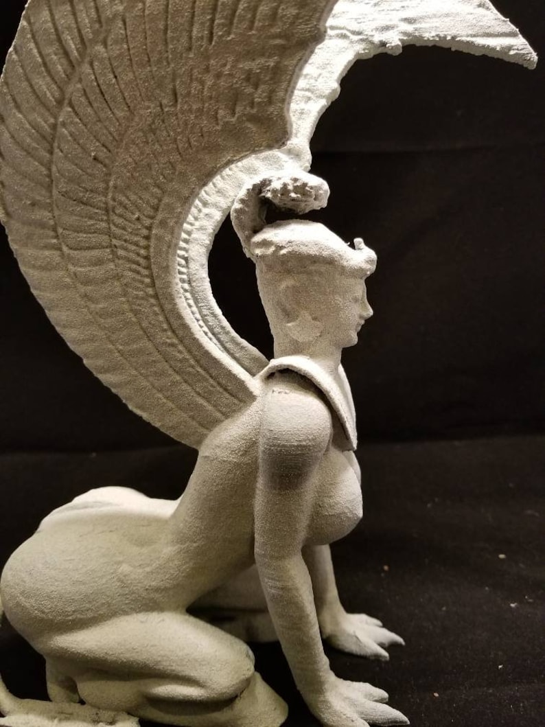 May include: A white, 3D printed sculpture of a winged woman with a crown, kneeling on all fours. The sculpture is detailed and has a textured surface.