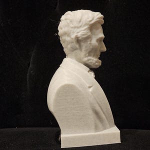 May include: A white plaster bust of a man with short, dark hair. The bust is on a white base and is viewed from the side.