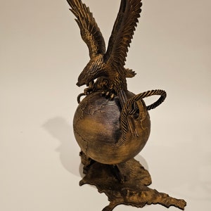 May include: A gold-colored eagle sculpture with its wings spread, perched atop a globe with a map of the world. The globe is on a stand shaped like the outline of the United States.