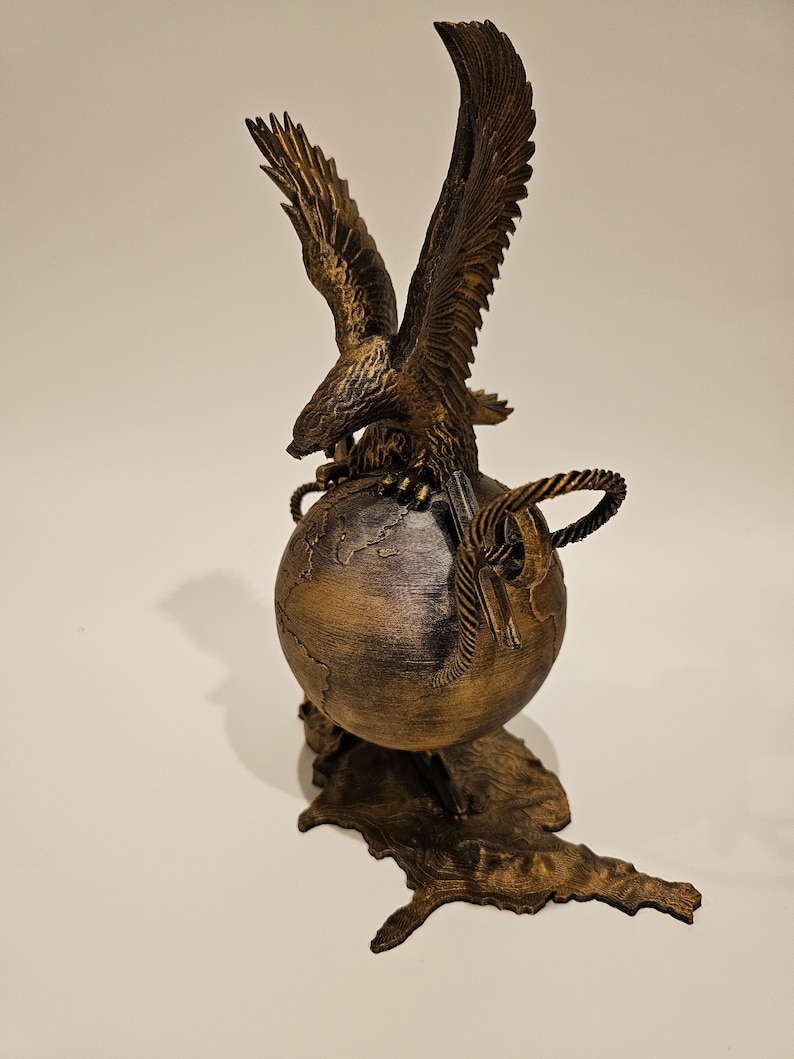 May include: A gold-colored eagle sculpture with its wings spread, perched atop a globe with a map of the world. The globe is on a stand shaped like the outline of the United States.