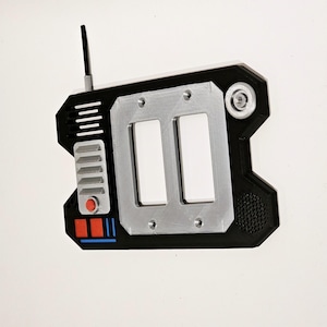 May include: A black and silver handheld device with a red button, a speaker, and a small antenna. The device has two rectangular slots and a textured surface.