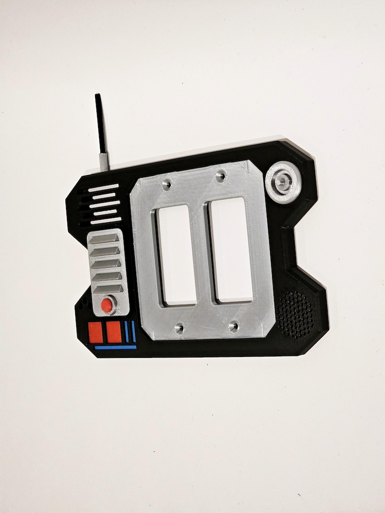 May include: A black and silver handheld device with a red button, a speaker, and a small antenna. The device has two rectangular slots and a textured surface.