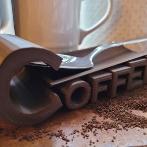 May include: A brown 3D printed coffee spoon holder with the word "COFFEE" printed on it. A silver spoon is resting on the holder.