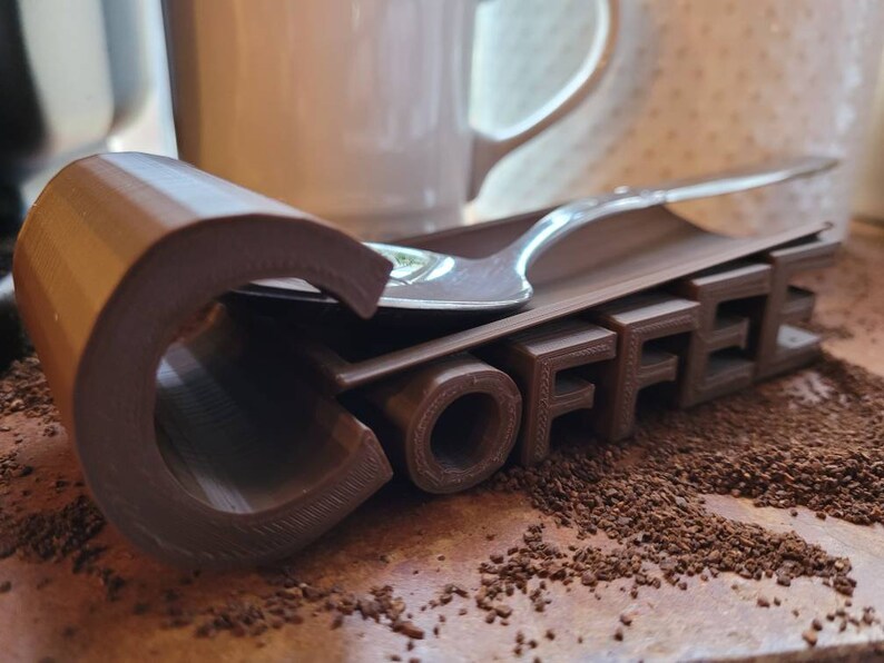 May include: A brown 3D printed coffee spoon holder with the word "COFFEE" printed on it. A silver spoon is resting on the holder.