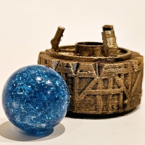 May include: A bright blue, spherical glass marble with a crackled, reflective interior texture positioned in front of a circular, metallic bronze-coloured object featuring raised geometric patterns and three small vertical protrusions on top.