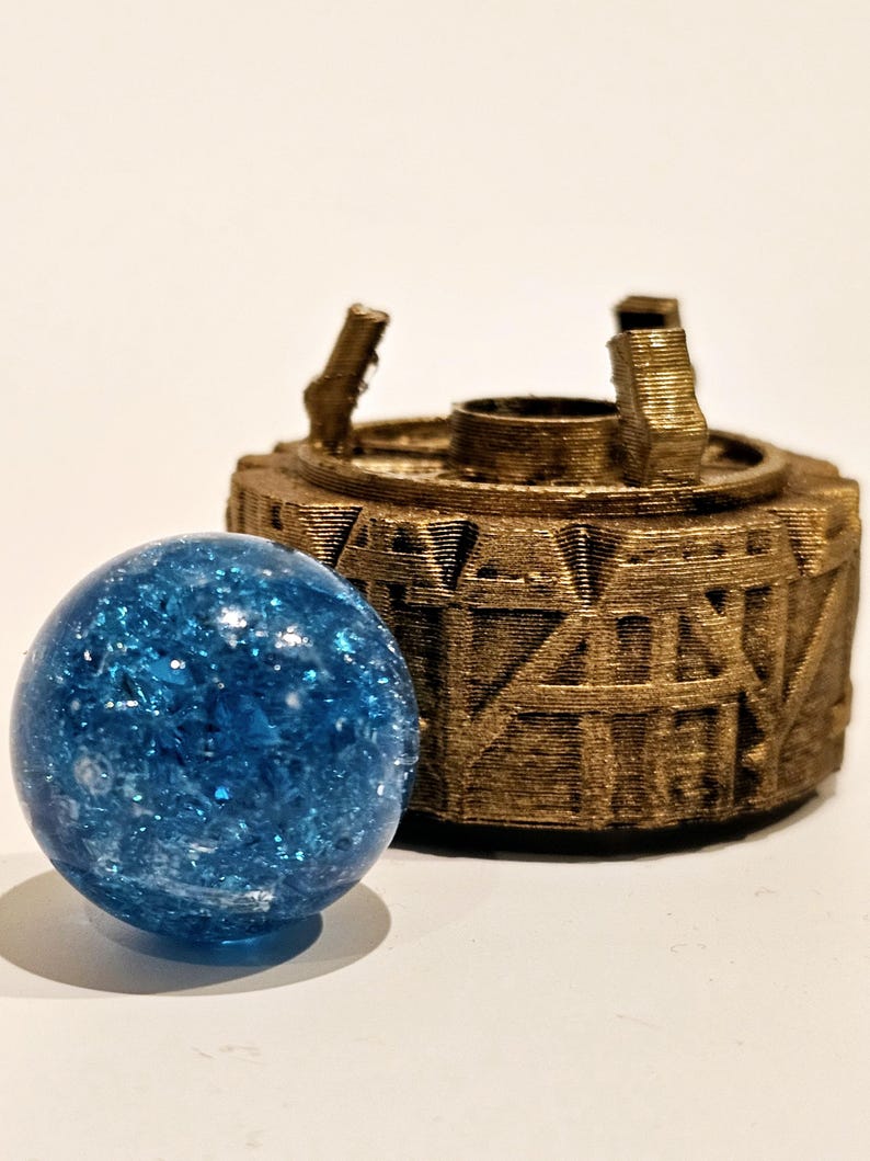 May include: A bright blue, spherical glass marble with a crackled, reflective interior texture positioned in front of a circular, metallic bronze-coloured object featuring raised geometric patterns and three small vertical protrusions on top.
