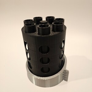 May include: A black plastic 3D printed cylinder with a circular base and a series of holes around the sides. The cylinder has seven round openings at the top, and a gray plastic base with a textured pattern.