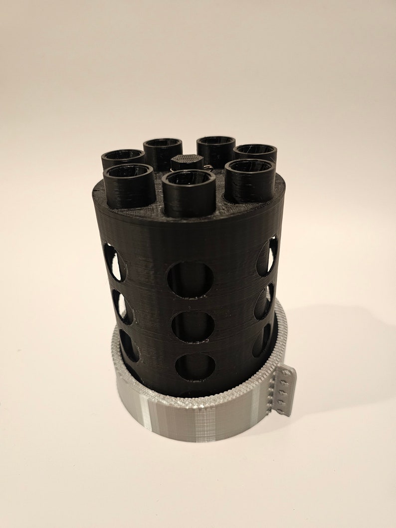 May include: A black plastic 3D printed cylinder with a circular base and a series of holes around the sides. The cylinder has seven round openings at the top, and a gray plastic base with a textured pattern.