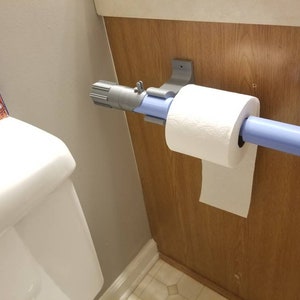 May include: A novelty toilet paper holder designed to resemble a lightsaber. The holder is light blue and gray, with a roll of white toilet paper in place. It is mounted on a wooden wall.