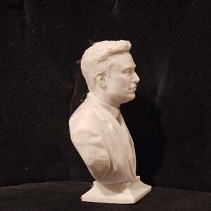 May include: A white bust sculpture of a person with short, dark hair. The sculpture is wearing a suit jacket and tie. The bust is on a white base.
