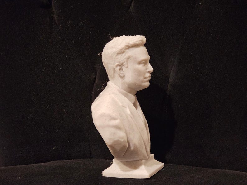 May include: A white bust sculpture of a person with short, dark hair. The sculpture is wearing a suit jacket and tie. The bust is on a white base.