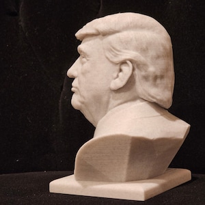 May include: A white marble bust sculpture of a person with short, wavy hair. The bust is on a white rectangular base.