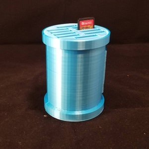 May include: A light blue plastic storage container with a gridded top for holding Nintendo Switch game cartridges. A game cartridge is inserted into the top of the container.