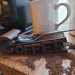 May include: A brown 3D printed coffee spoon holder with the word "COFFEE" printed on it. A silver spoon is resting on top of the holder.