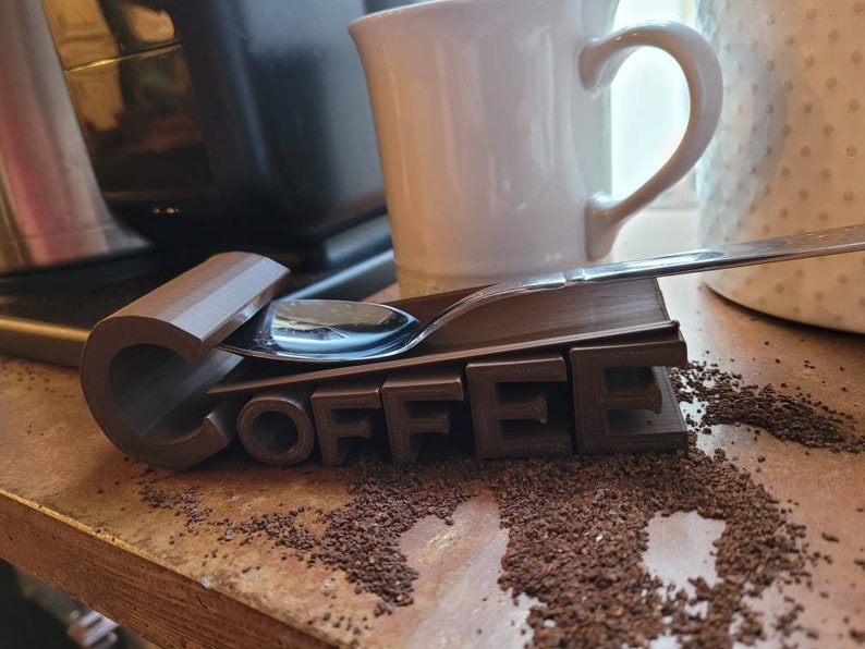 May include: A brown 3D printed coffee spoon holder with the word "COFFEE" printed on it. A silver spoon is resting on top of the holder.