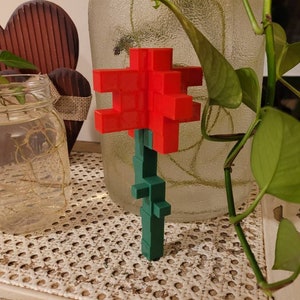 May include: A red and green 3D printed flower made of small blocks. The flower is sitting on a table with a woven pattern.