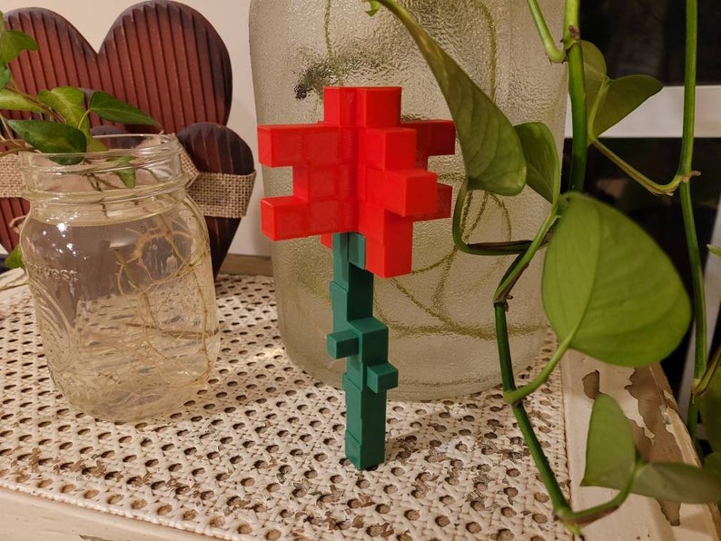 May include: A red and green 3D printed flower made of small blocks. The flower is sitting on a table with a woven pattern.