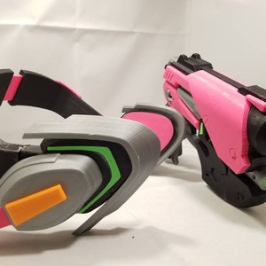 Puede incluir: Una pistola impresa en 3D de color rosa y negro y un auricular impreso en 3D de color gris y verde. El auricular tiene un acento naranja.