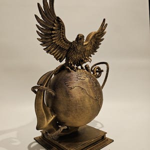 May include: A bronze-colored sculpture of an eagle with outstretched wings perched atop a globe. The globe is wrapped with a rope and sits on a square base. The sculpture has intricate details and a weathered finish.