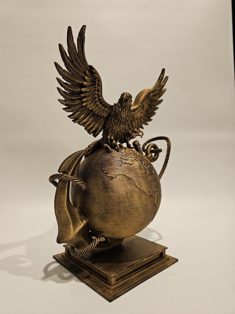 May include: A bronze-colored sculpture of an eagle with outstretched wings perched atop a globe. The globe is wrapped with a rope and sits on a square base. The sculpture has intricate details and a weathered finish.