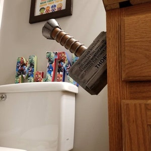 May include: A wooden replica of Thor's hammer, Mjolnir, with the inscription "Whosoever sits here and holds this hammer, is deemed worthy and possesses the power of Thor." The hammer is mounted on a wall next to a white toilet and a framed print with the text "Even superheroes take a bath, flush the toilet, wash their hands, brush their teeth."