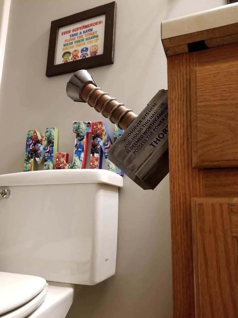 May include: A wooden replica of Thor's hammer, Mjolnir, with the inscription "Whosoever sits here and holds this hammer, is deemed worthy and possesses the power of Thor." The hammer is mounted on a wall next to a white toilet and a framed print with the text "Even superheroes take a bath, flush the toilet, wash their hands, brush their teeth."
