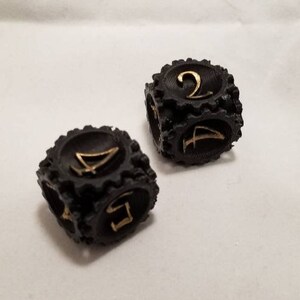 May include: Two black dice with gold numbers. The dice have a textured, tire-like design. One die shows the number 2 and the other die shows the number 4.