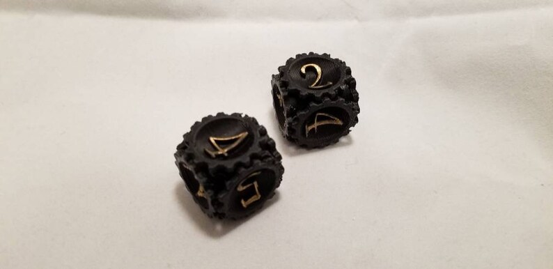 May include: Two black dice with gold numbers. The dice have a textured, tire-like design. One die shows the number 2 and the other die shows the number 4.