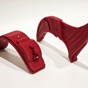 Op de afbeelding: Twee 3D-geprinte rode kunststof onderdelen voor een project. Een onderdeel is een gebogen stuk met een kleine knop en een gleuf. Het andere onderdeel is een gebogen stuk met een complexer ontwerp.