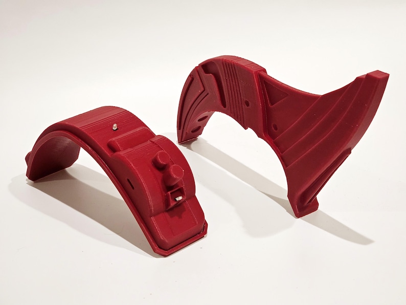 Op de afbeelding: Twee 3D-geprinte rode kunststof onderdelen voor een project. Een onderdeel is een gebogen stuk met een kleine knop en een gleuf. Het andere onderdeel is een gebogen stuk met een complexer ontwerp.