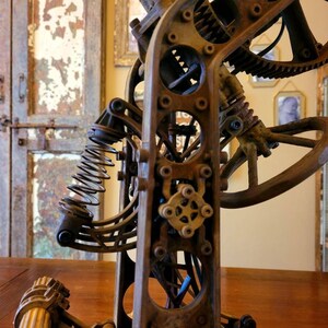 Puede incluir: Primer plano de una escultura de metal oxidado con engranajes, un resorte y un mango de madera. La escultura est&aacute; hecha de varias piezas de metal y tiene una est&eacute;tica steampunk.