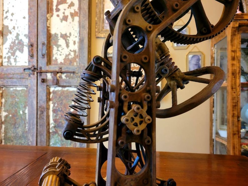 Puede incluir: Primer plano de una escultura de metal oxidado con engranajes, un resorte y un mango de madera. La escultura est&aacute; hecha de varias piezas de metal y tiene una est&eacute;tica steampunk.