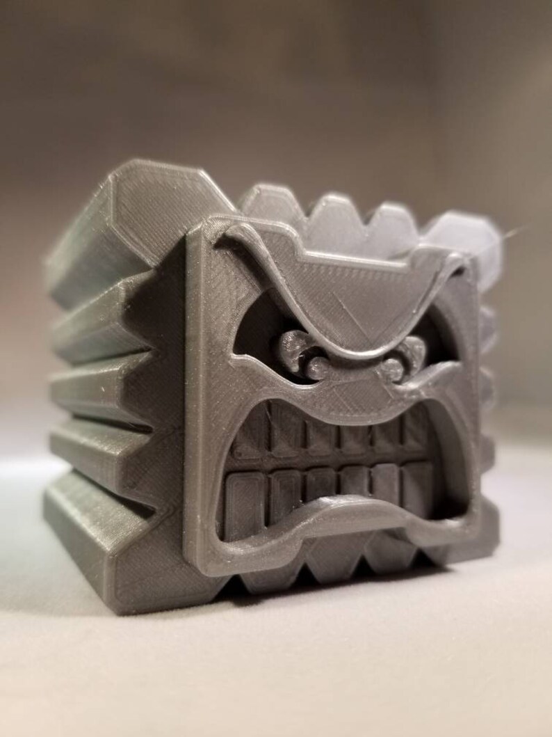 K&ouml;nnte beinhalten: Ein grauer 3D-gedruckter Block mit einem bedrohlichen Gesicht. Der Block hat eine strukturierte Oberfl&auml;che und eine Reihe von Z&auml;hnen um den Mund.