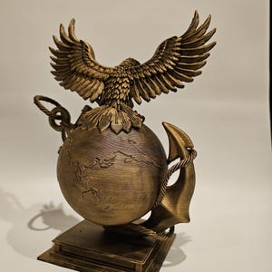 May include: A bronze-colored sculpture featuring an eagle with outstretched wings atop a globe. An anchor and rope detail the globe's side. The sculpture is mounted on a square base, showcasing intricate details and textures.