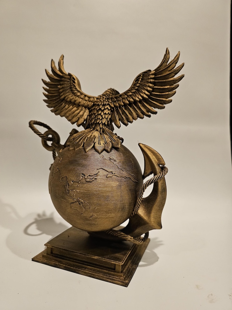 May include: A bronze-colored sculpture featuring an eagle with outstretched wings atop a globe. An anchor and rope detail the globe's side. The sculpture is mounted on a square base, showcasing intricate details and textures.