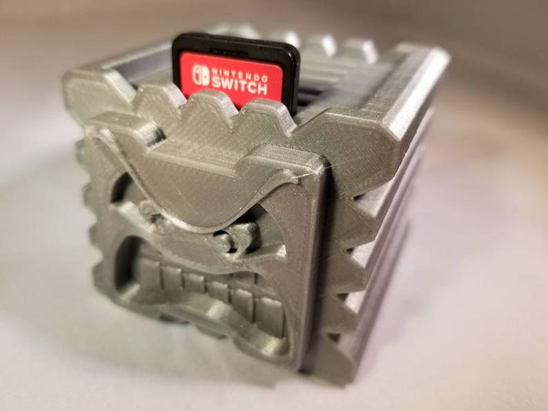 K&ouml;nnte beinhalten: Ein grauer, 3D-gedruckter Spielkartuschenhalter in Form eines Monstergesichts. Der Halter hat einen Schlitz f&uuml;r eine Nintendo Switch-Spielkartusche. Die Kartusche wird in den Halter eingesetzt und tr&auml;gt die Aufschrift "Nintendo Switch".