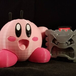 K&ouml;nnte beinhalten: Eine rosa und graue 3D-gedruckte Figur eines Videospielcharakters, Kirby, steht neben einem grauen Block mit einem bedrohlichen Gesicht.