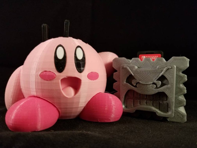 K&ouml;nnte beinhalten: Eine rosa und graue 3D-gedruckte Figur eines Videospielcharakters, Kirby, steht neben einem grauen Block mit einem bedrohlichen Gesicht.