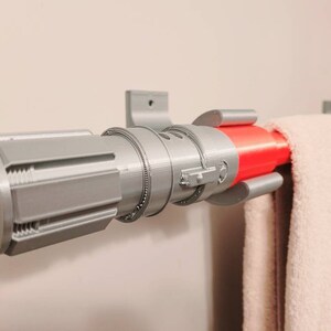 May include: A silver lightsaber-shaped towel rack with a red accent. The rack is mounted to a wall and has a towel hanging from it.