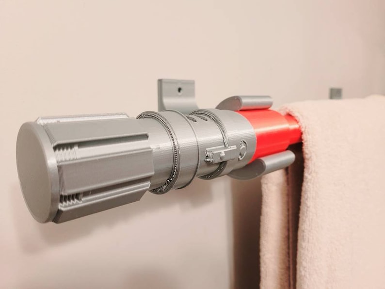 May include: A silver lightsaber-shaped towel rack with a red accent. The rack is mounted to a wall and has a towel hanging from it.
