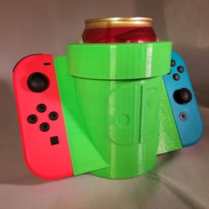 Op de afbeelding: Een groene 3D-geprinte drankhouder in de vorm van een videogamepijp, met aan weerszijden een rode en blauwe videogamecontroller. Er zit een frisdrankblikje in de houder. De drankhouder is ontworpen om een blikje en twee controllers vast te houden.