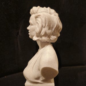 May include: A white bust sculpture of a woman with short, wavy hair. The sculpture is viewed from the side, showing the woman's profile.
