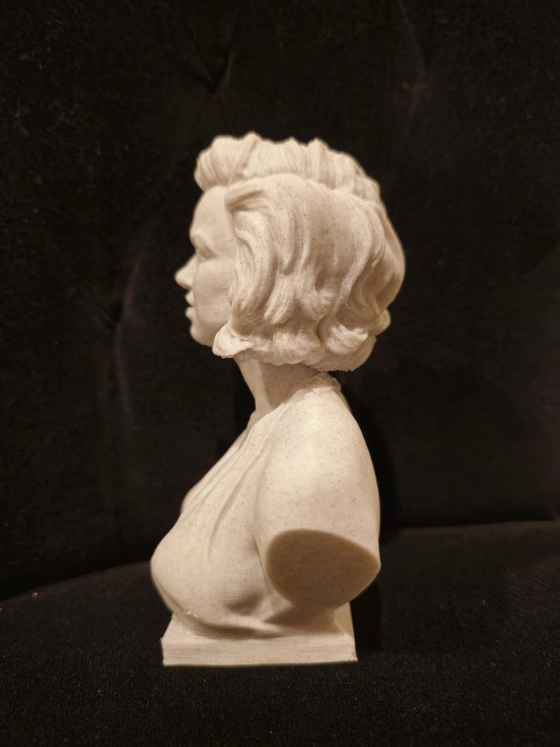 May include: A white bust sculpture of a woman with short, wavy hair. The sculpture is viewed from the side, showing the woman's profile.