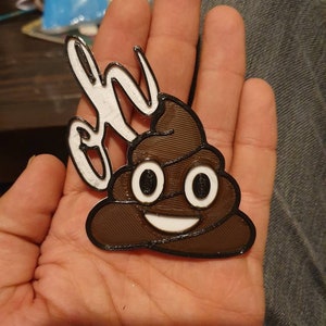 May include: A 3D printed brown poop emoji with a white smiling face and eyes. The emoji is attached to a white 3D printed object that says "Oh" in white.