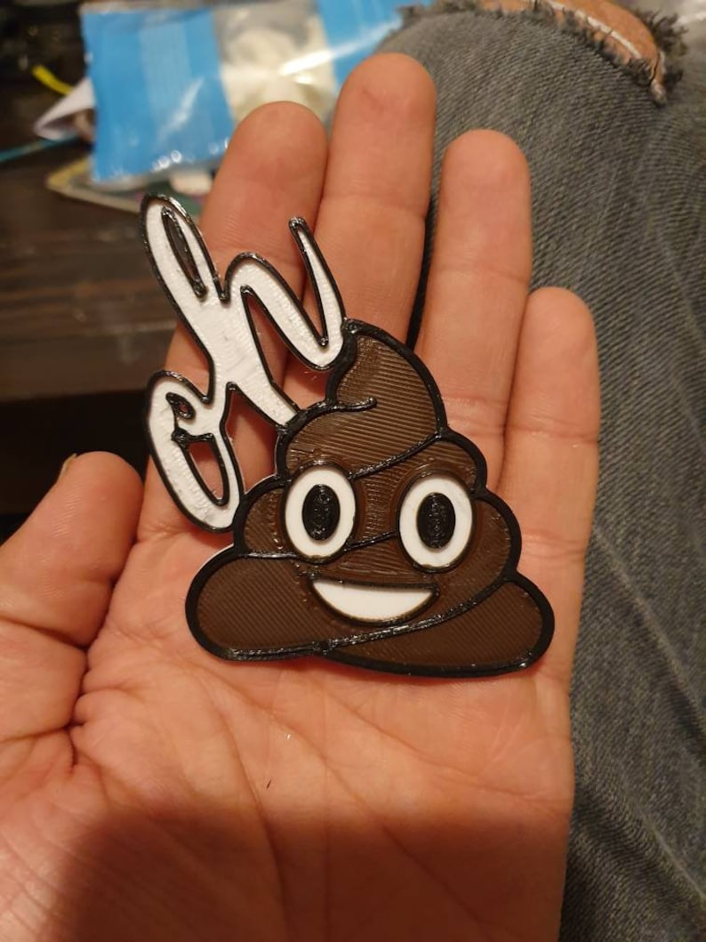 May include: A 3D printed brown poop emoji with a white smiling face and eyes. The emoji is attached to a white 3D printed object that says "Oh" in white.